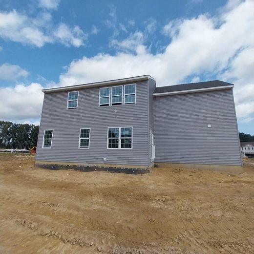 Two-story house under construction with gray siding against a blue sky, dirt ground.