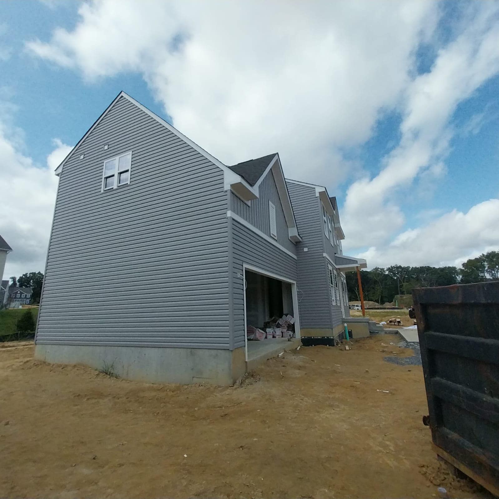 New house under construction, gray siding, blue sky, empty lot, a dumpster.