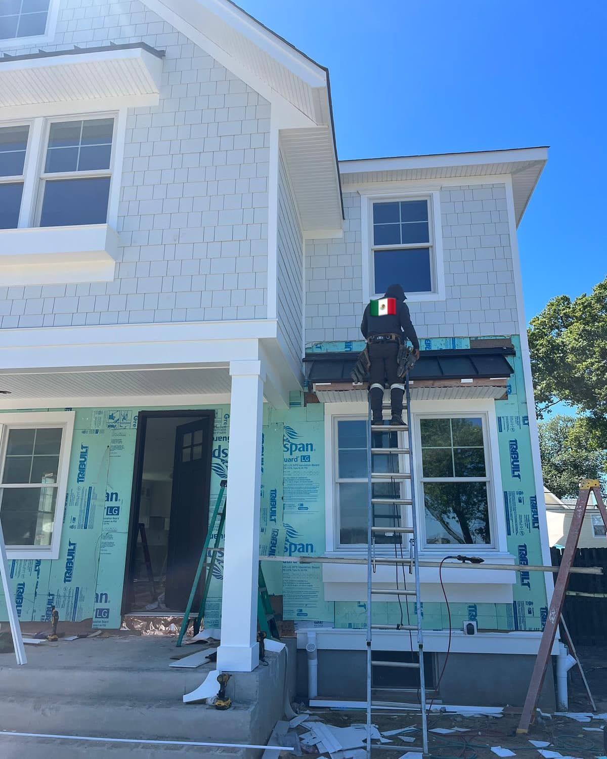 Construction worker on a ladder installing a roof, siding, and windows on a new light blue house on a sunny day.