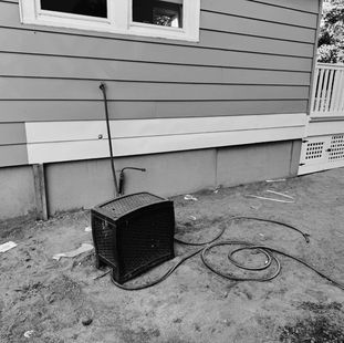 A black, boxy appliance sits on dirt next to a light-colored house wall with a window. Hoses are coiled nearby.