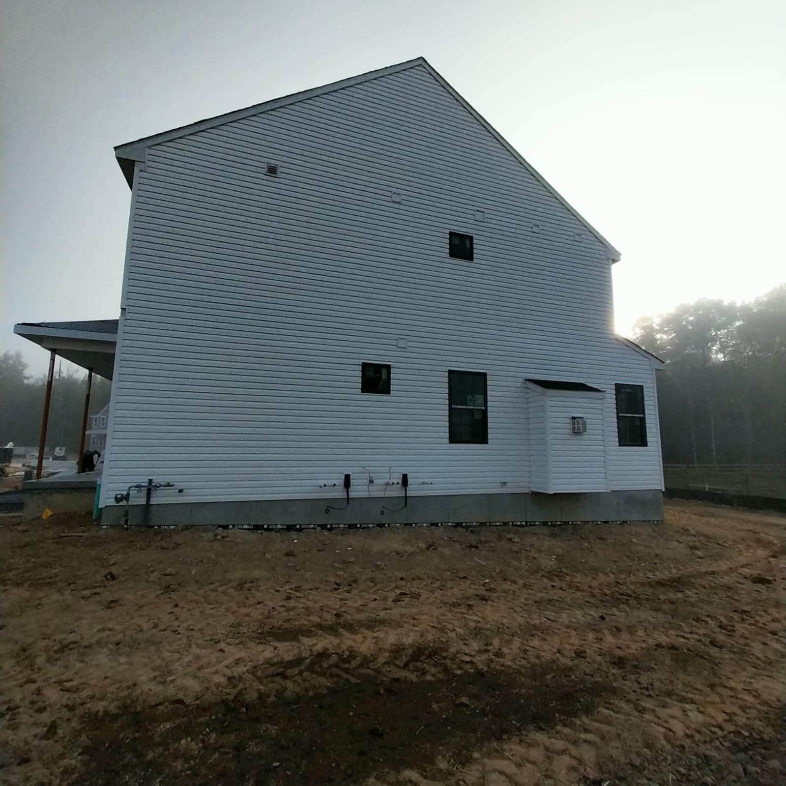 Side view of a two-story house with white siding under construction on a dirt lot, overcast sky.
