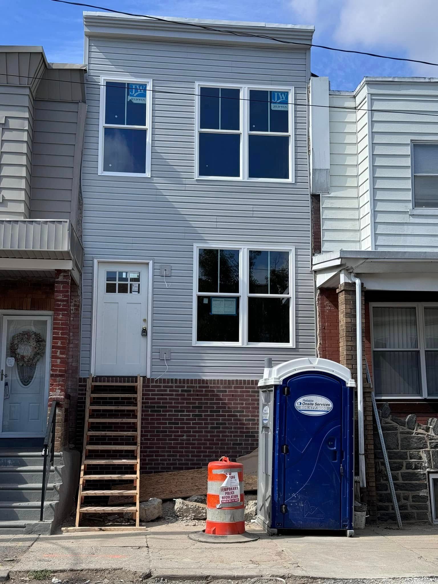 Gray two-story house under construction with white windows, a temporary staircase, and a portable toilet.