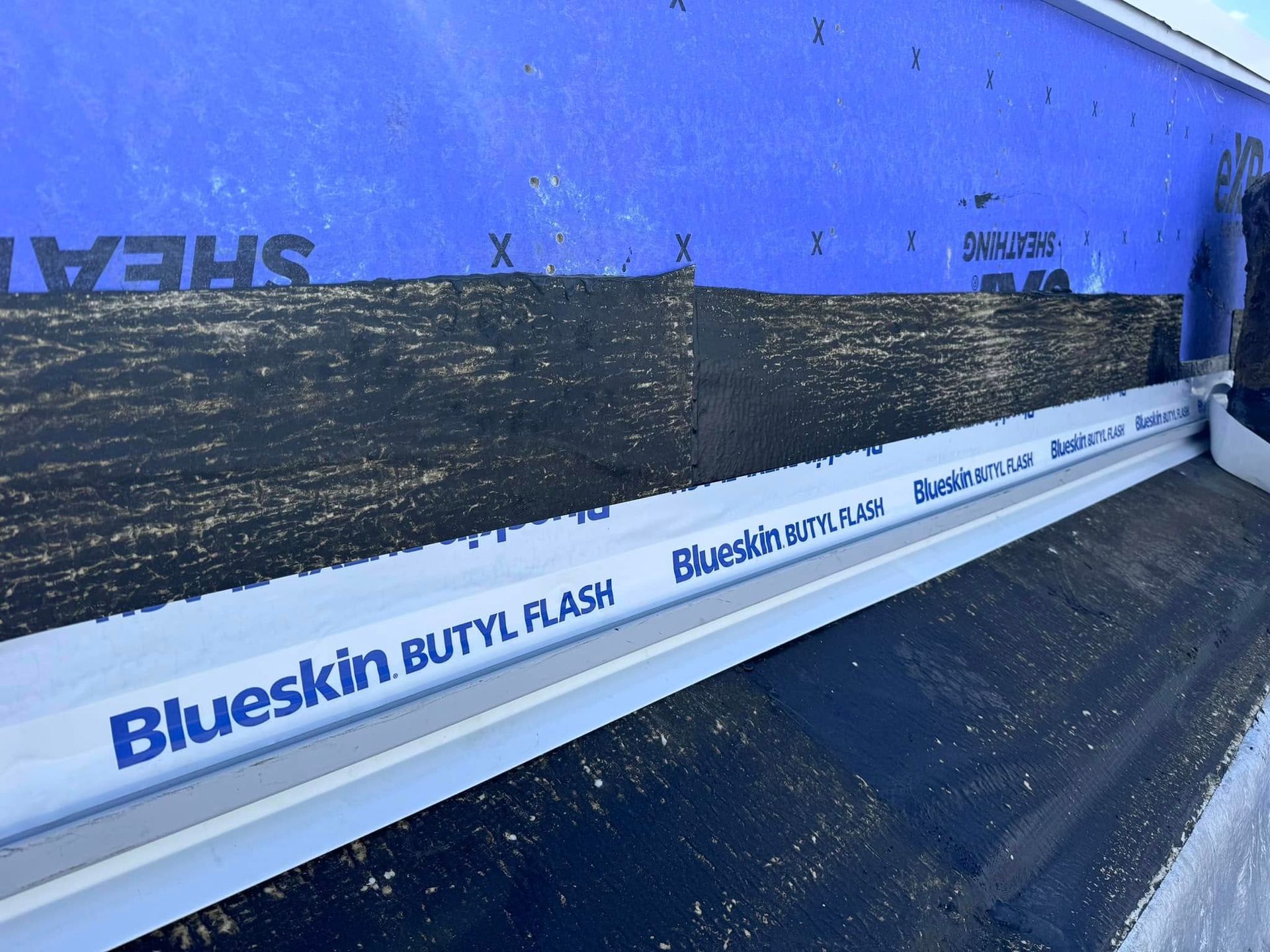 Blue flashing tape (Blueskin Butyl Flash) installed on a roof with blue underlayment and black sheathing.