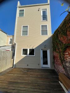 Three-story beige building with multiple windows and a back door, facing a wooden deck. A brick wall with vines is to the right.