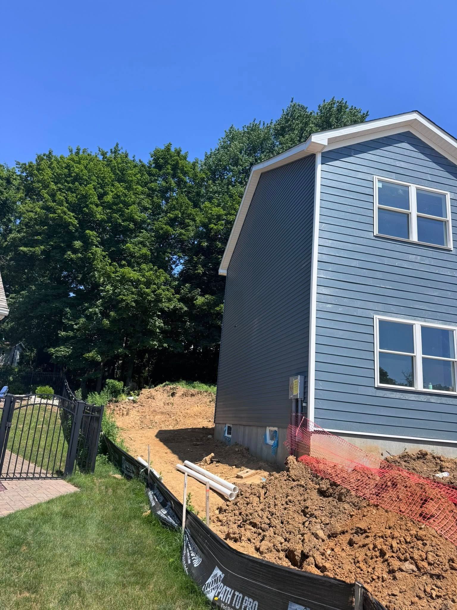 Blue-sided house beside a dirt embankment held by black landscape fabric under a blue sky, tall green trees.