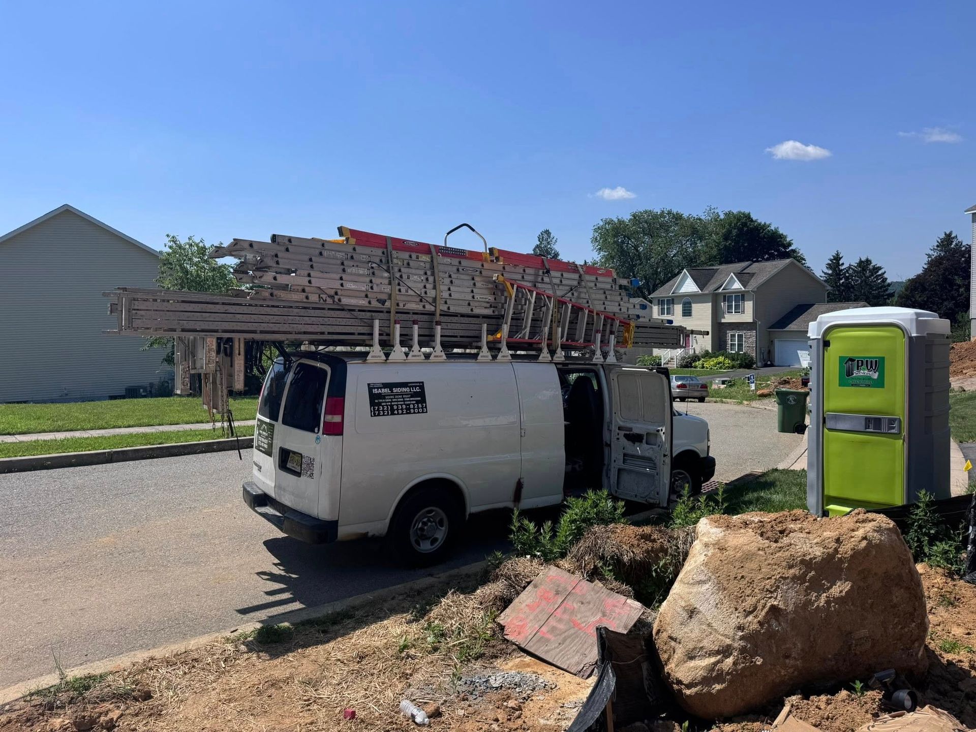 White work van with ladders on top, parked on a street near a construction site.
