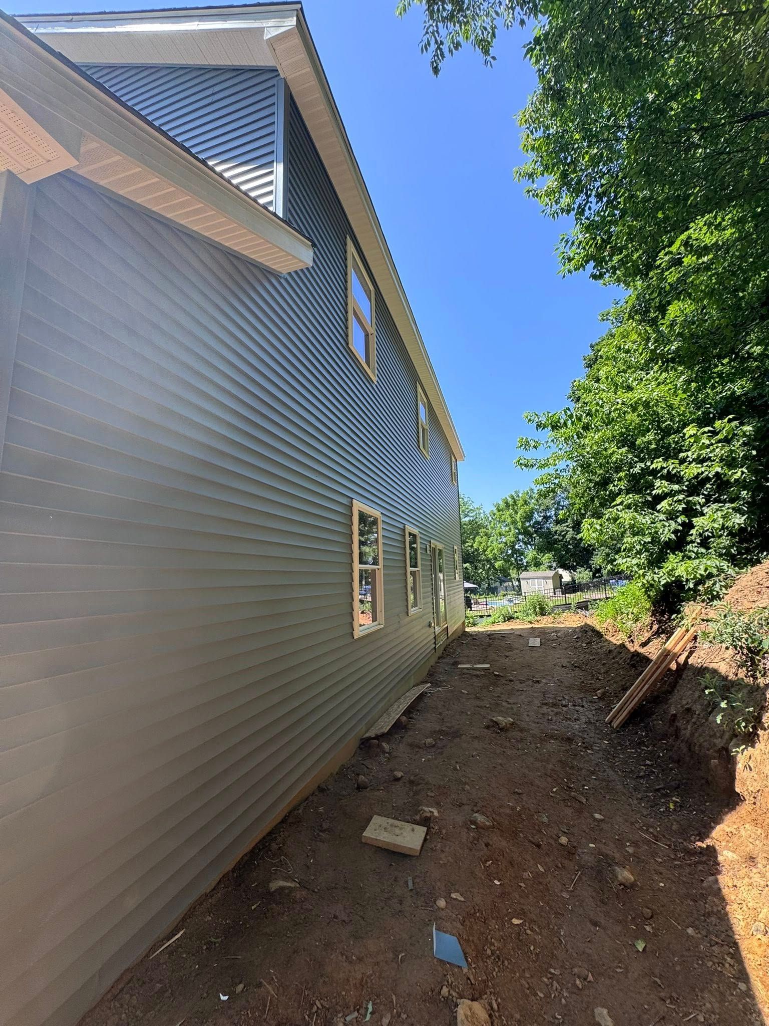 Side view of a two-story blue house with windows, next to a dirt path and green trees under a blue sky.