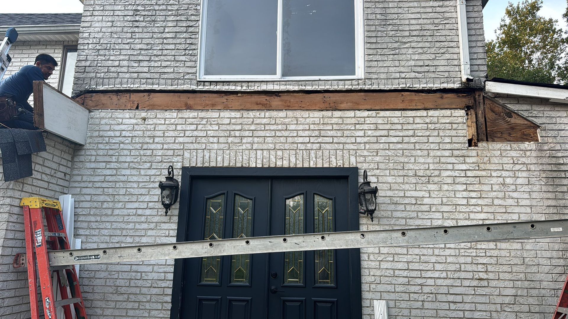 Man working on exterior brick wall renovation; wood beam exposed above front door and window.