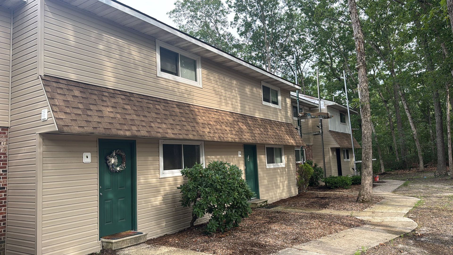 Two-story tan townhouses with brown roofs and green doors, set in a wooded area.