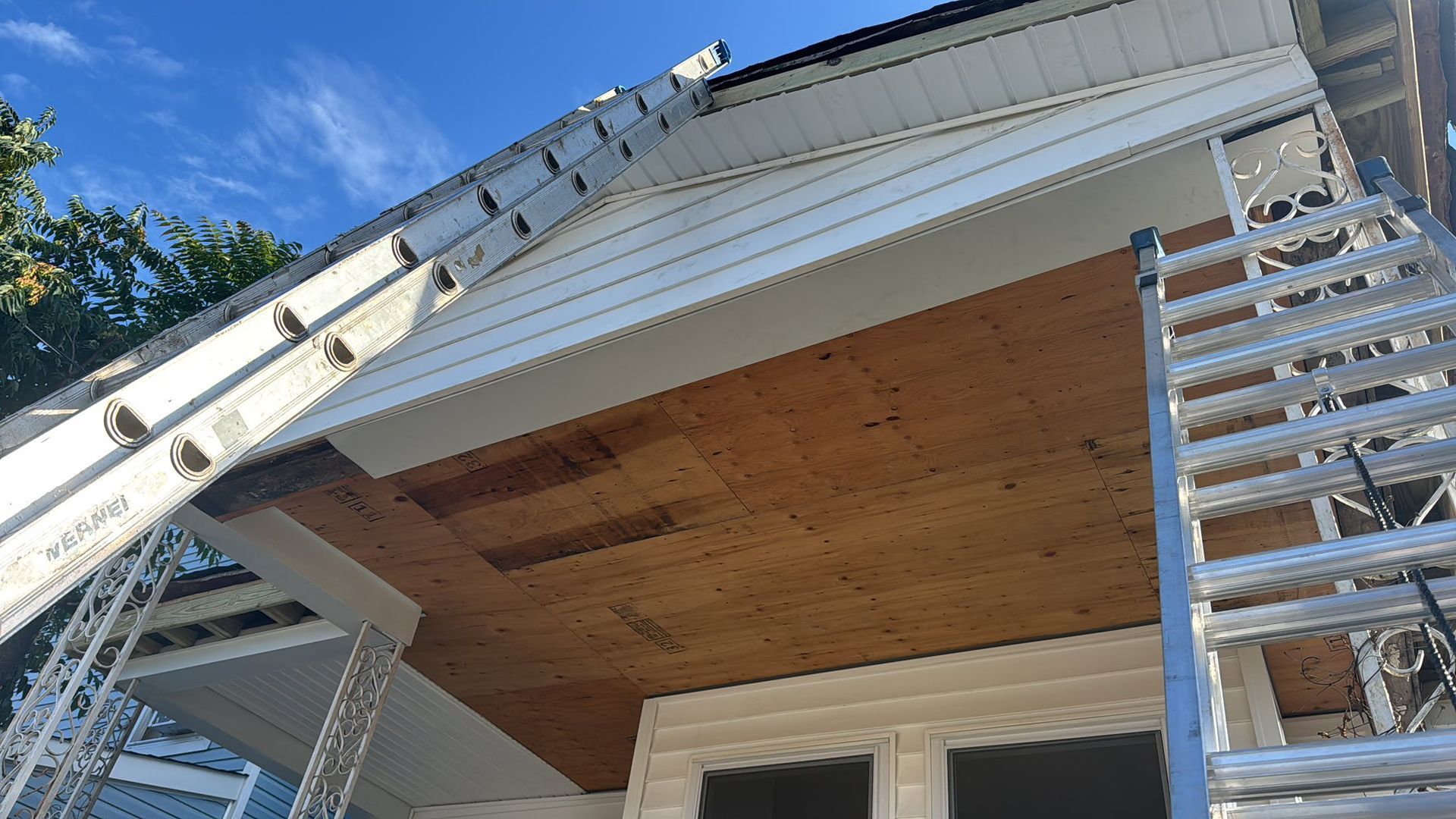 Ladders leaning against a building's underside, exposing plywood. New siding being installed on the upper portion.