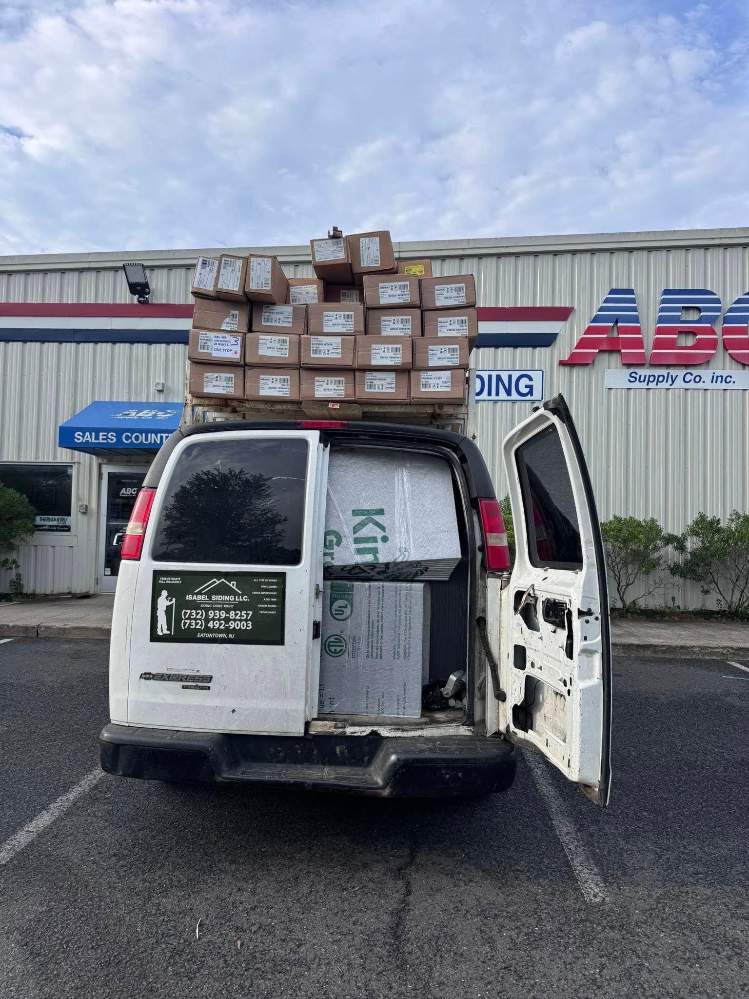 White van with open back, loaded with boxes on roof and inside, parked in front of a building.