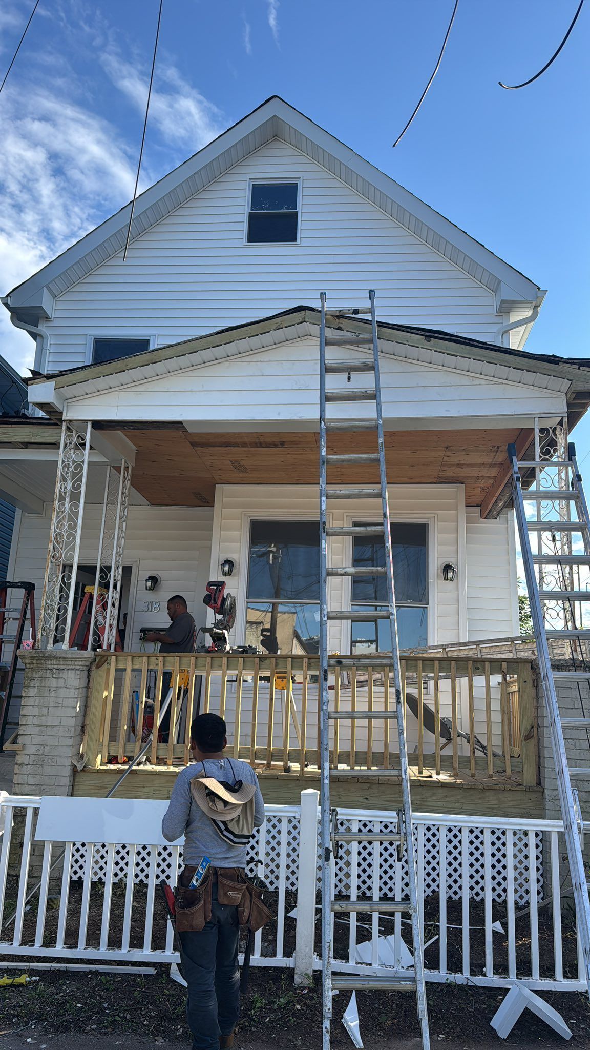 Workers renovating a white house with a porch, ladders, and scaffolding under a blue sky.