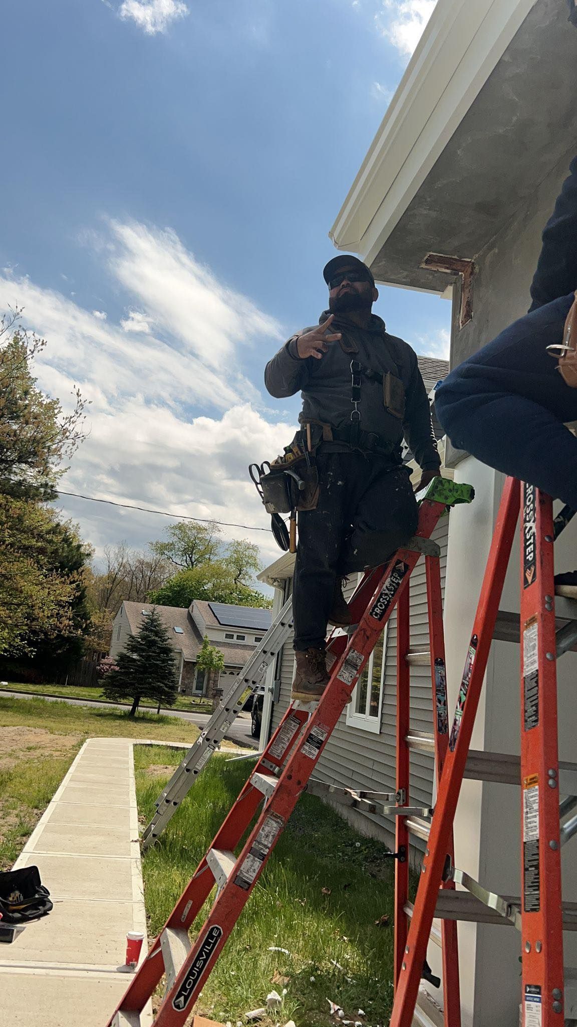 Man on ladder installing gutter, gesturing outdoors under cloudy sky.