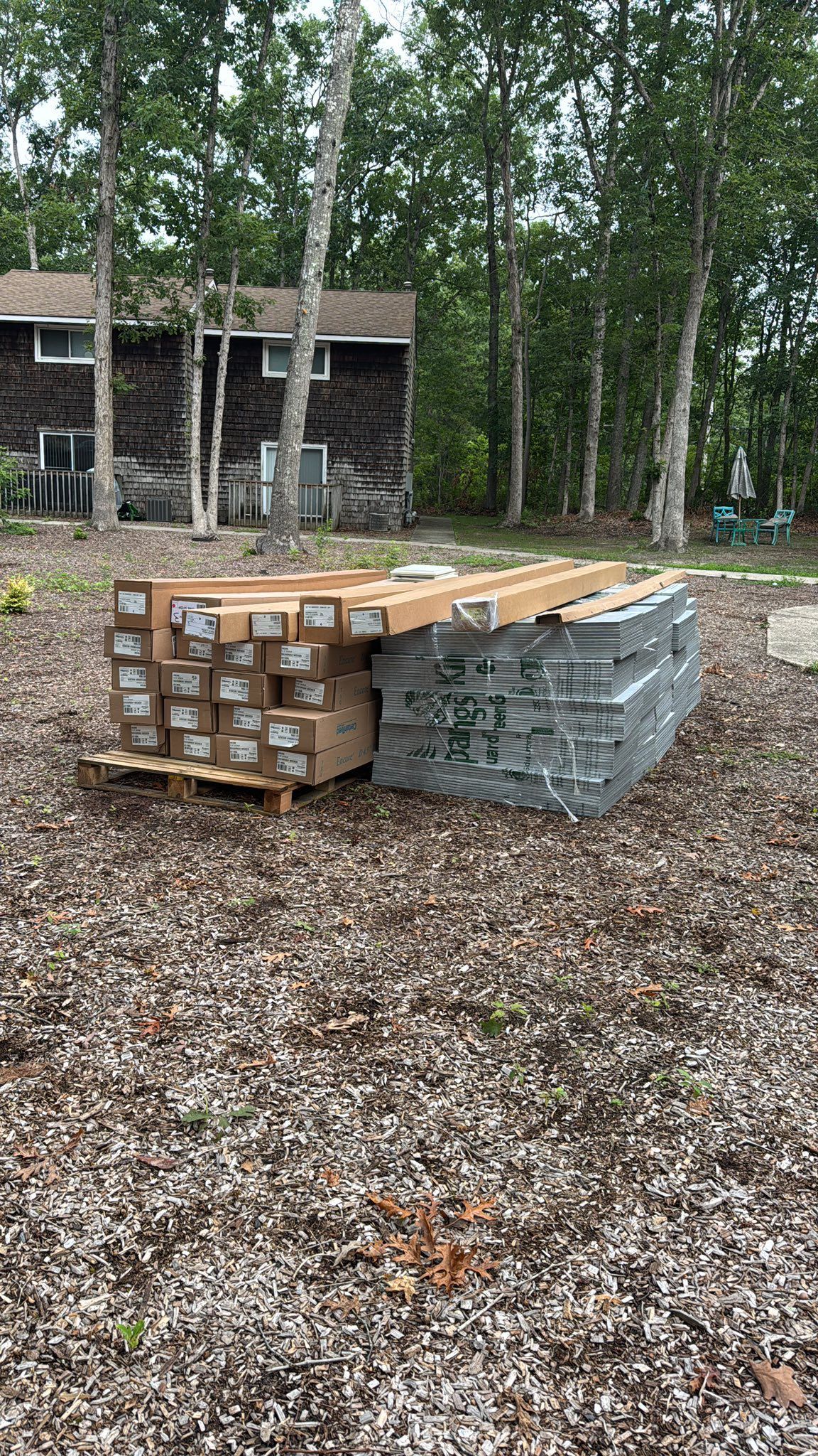 Two pallets with building materials sit on mulch in front of a house and trees.
