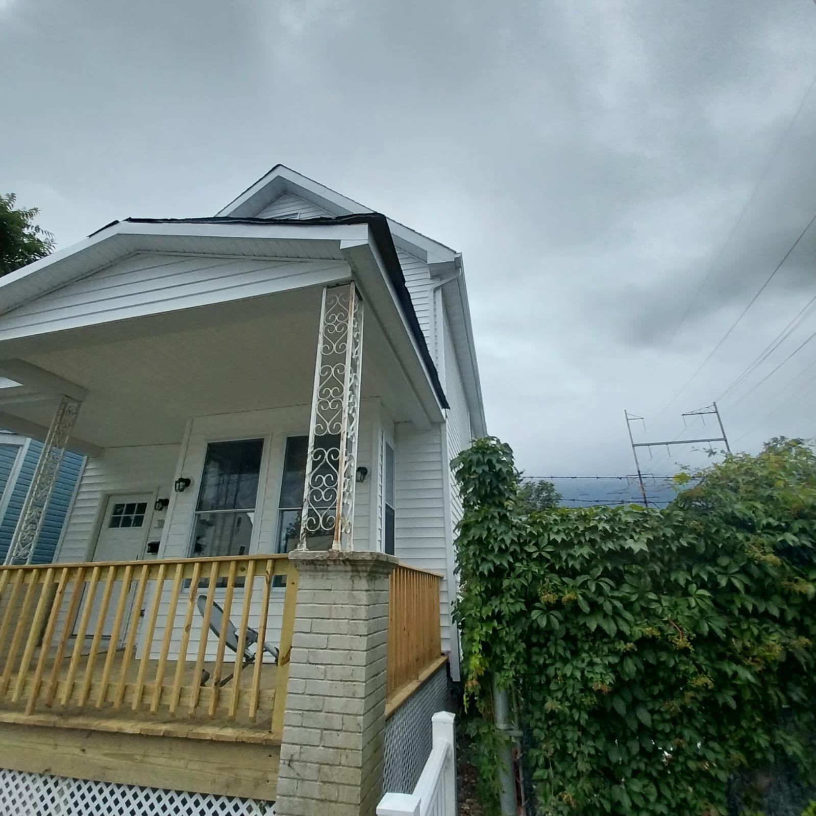 White house with porch under a cloudy sky. Brick and decorative posts support the porch roof. Green vines grow on the side.