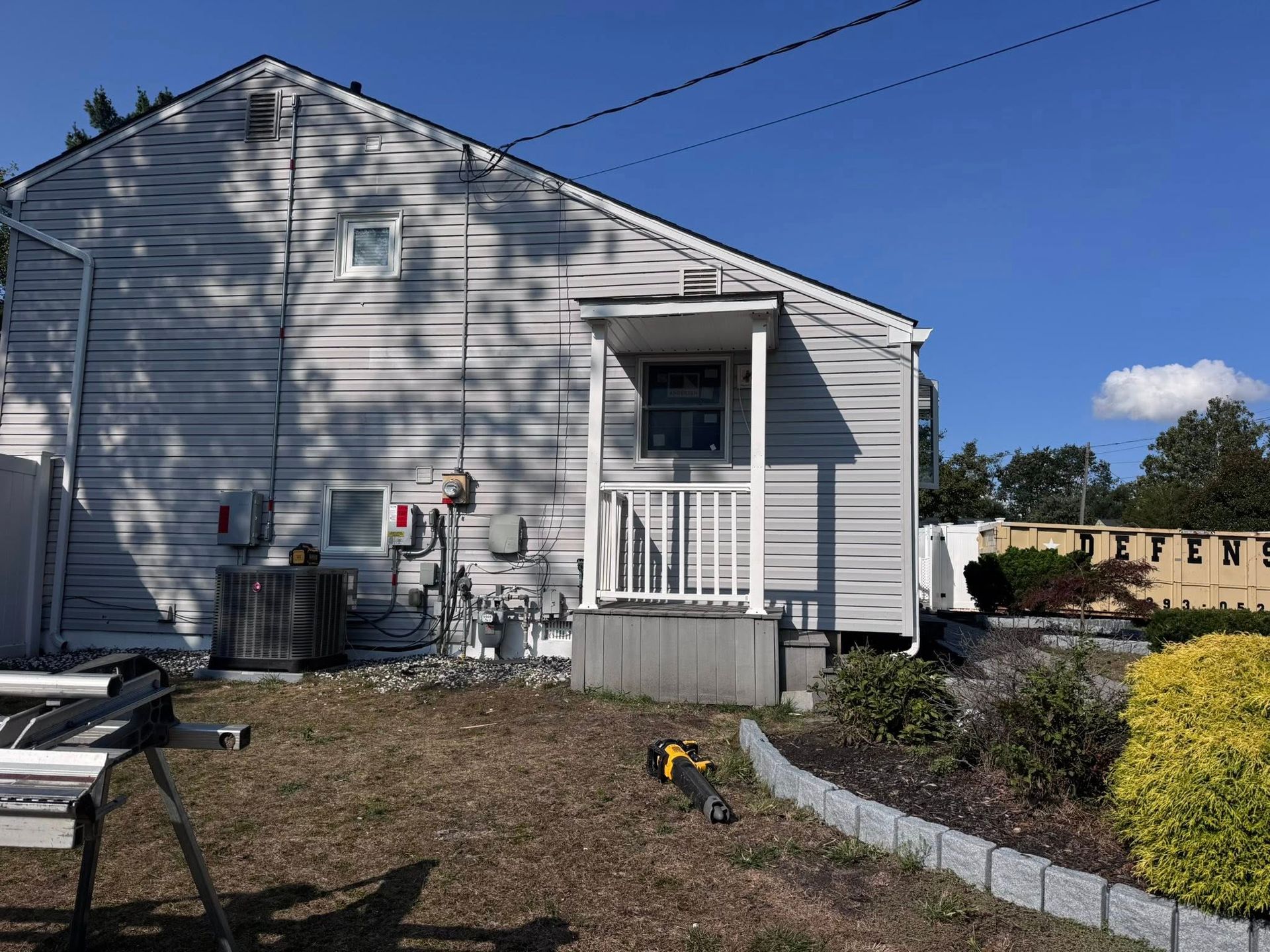 Side of a house with gray siding, a small porch, and an overgrown yard under a blue sky.