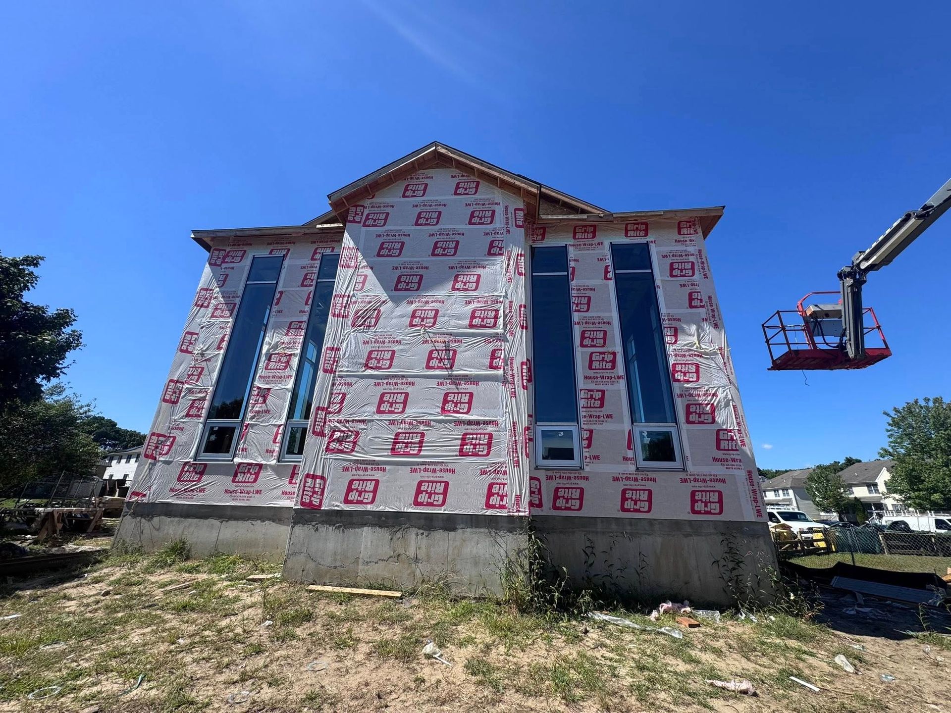 House under construction; exterior with tall windows, wrapped in red and white material, clear blue sky.