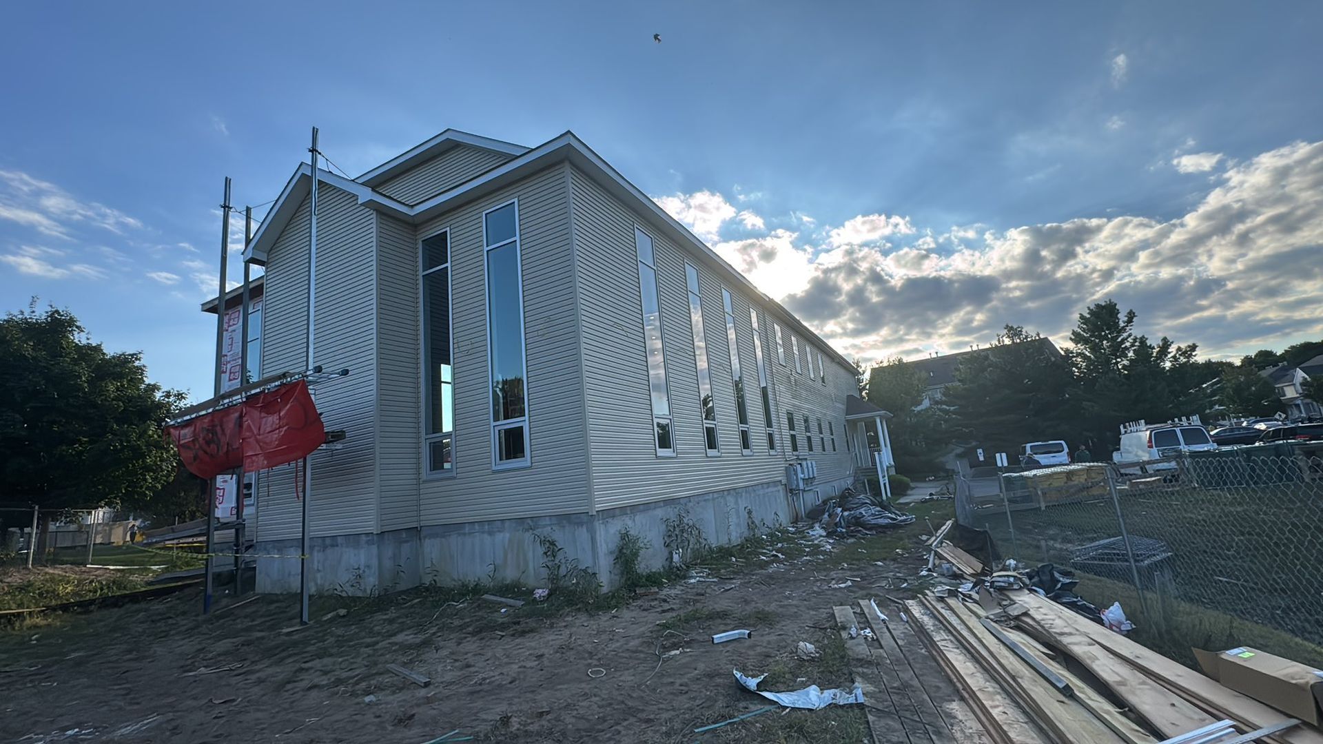 Building exterior under construction; light-colored siding, long windows, partially cloudy sky, dirt lot.