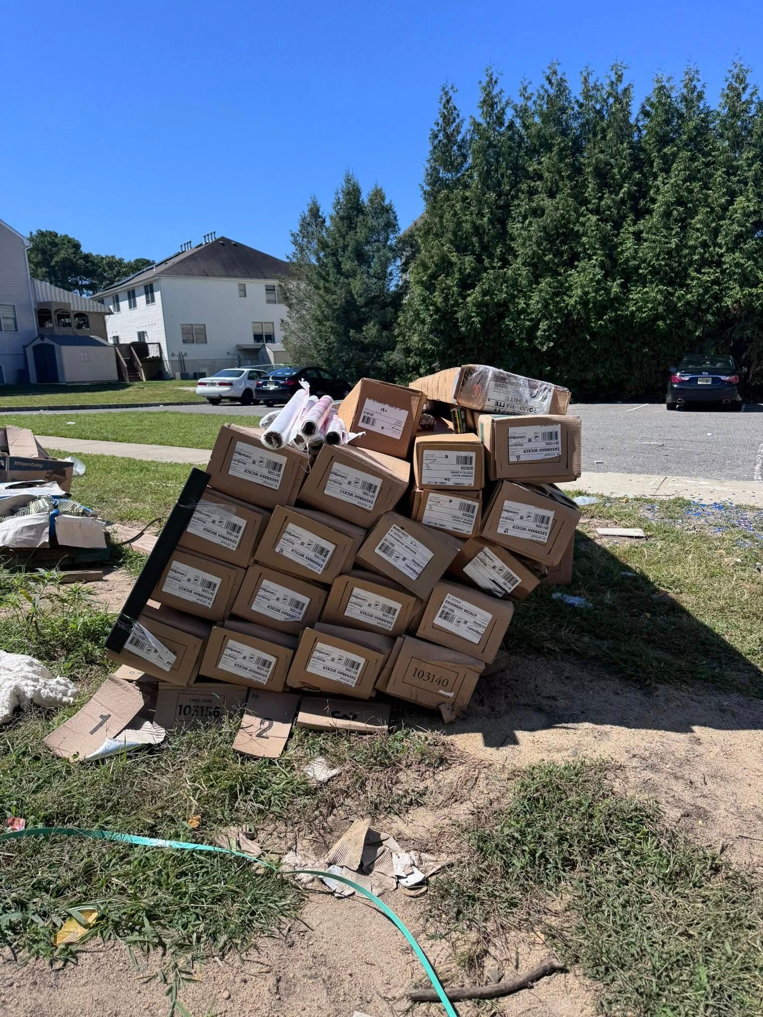 Pile of cardboard boxes stacked on grass near a residential street, under a blue sky.