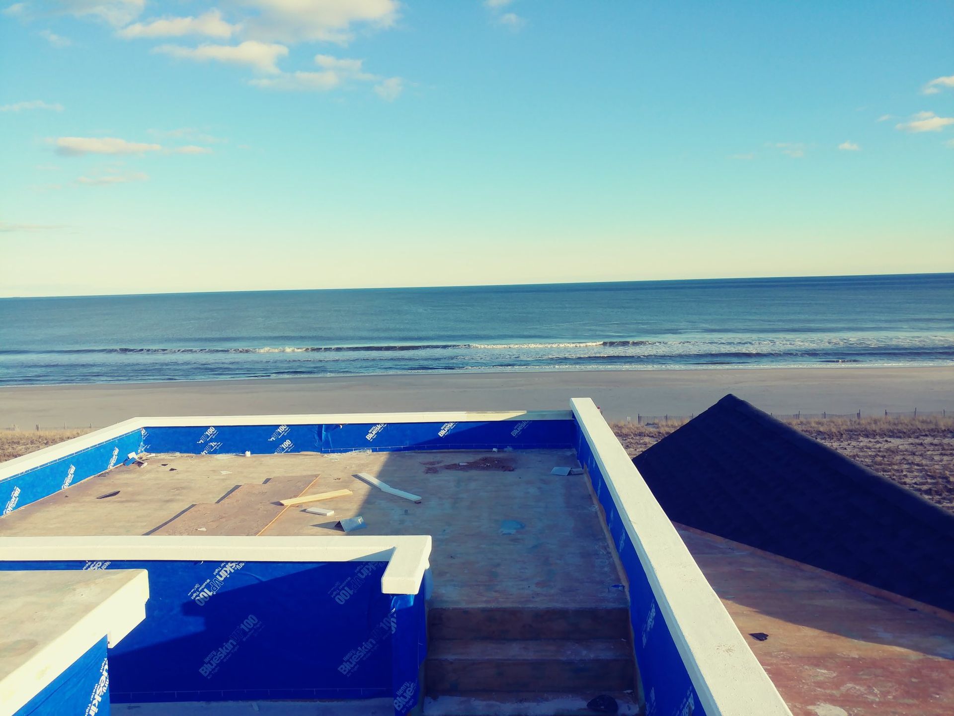 Rooftop with blue waterproofing, overlooking a beach with ocean waves under a blue sky.