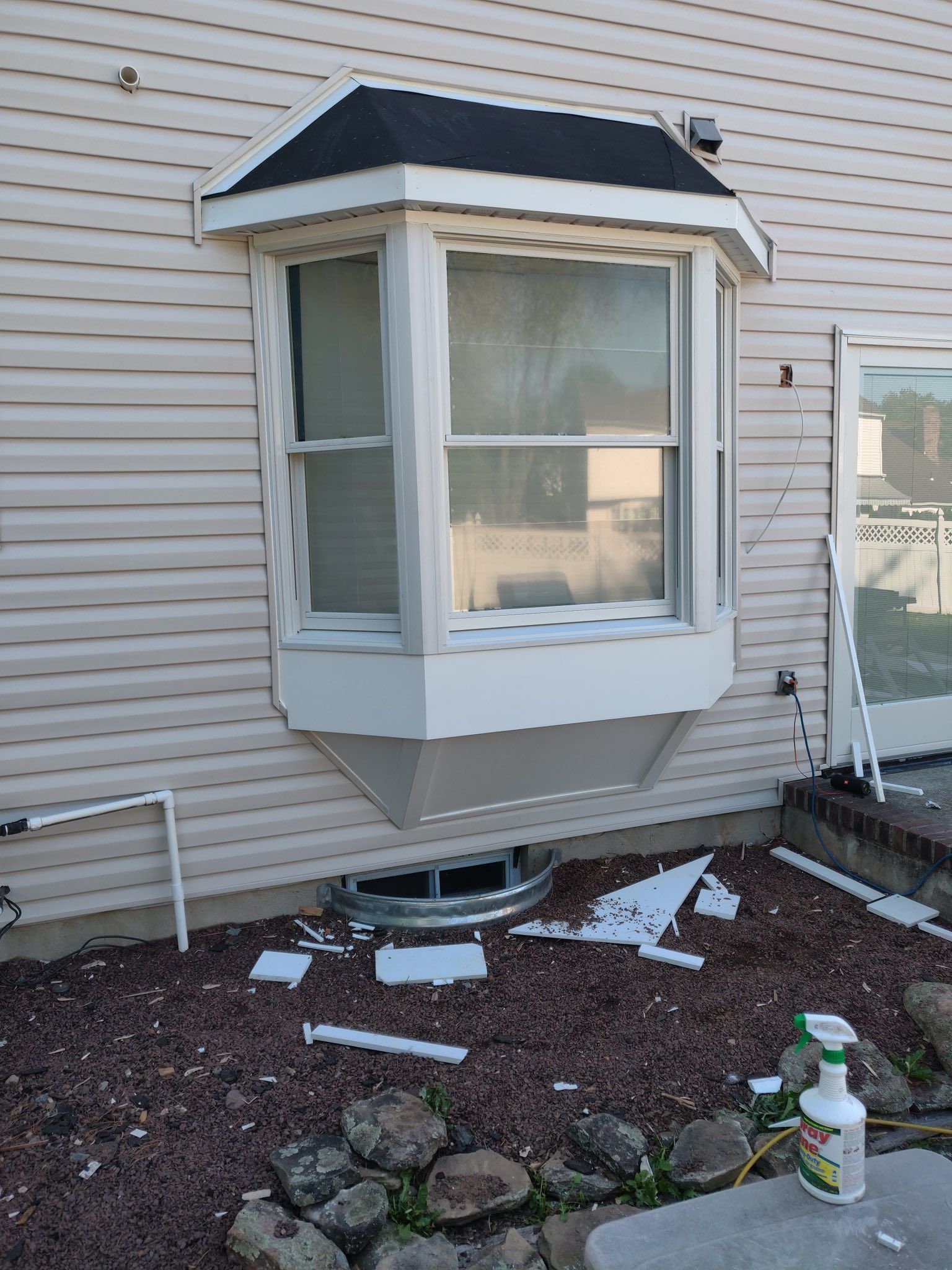 Bay window on a beige house; window well visible. Debris and rocks in the foreground.