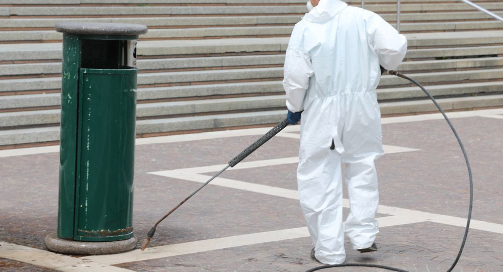 Person in white protective suit sprays pavement near a green trash can.