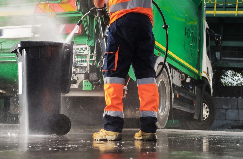 Worker power-washes a black trash bin near a green garbage truck; worker wears safety vest and orange-trimmed pants.