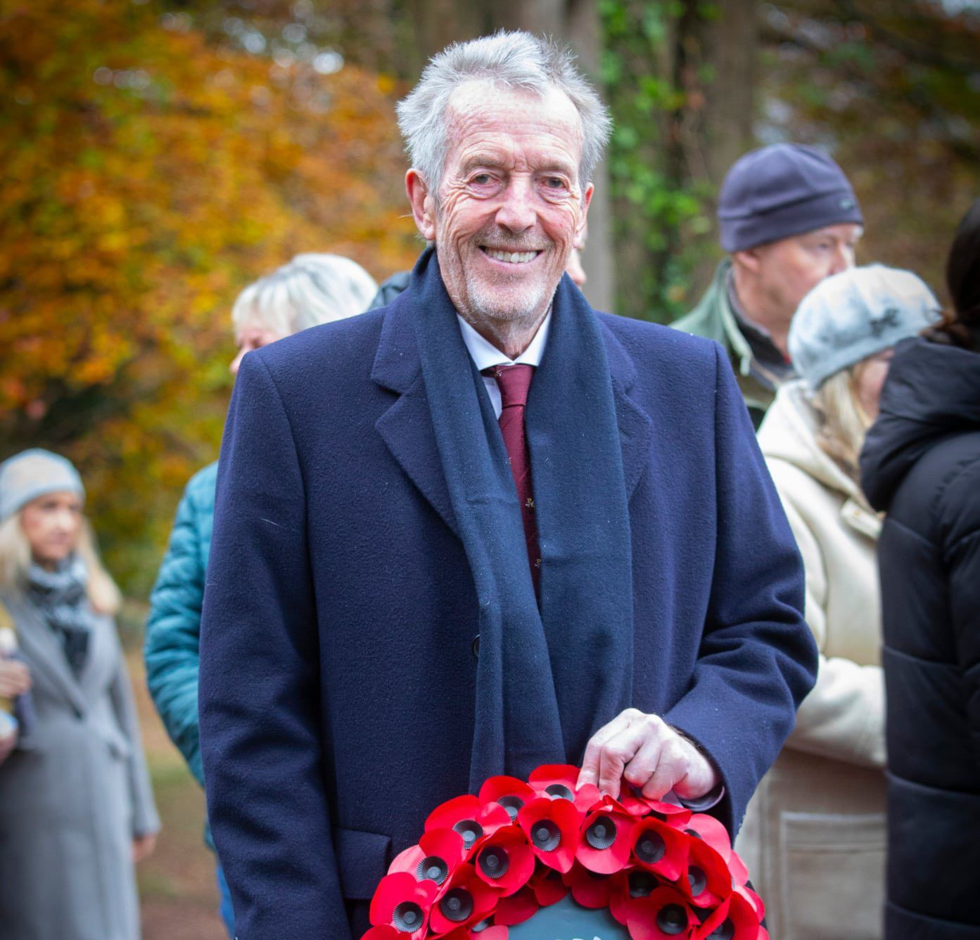 Man holding a poppy wreath outdoors, smiling. He wears a navy coat and scarf. Others stand nearby.