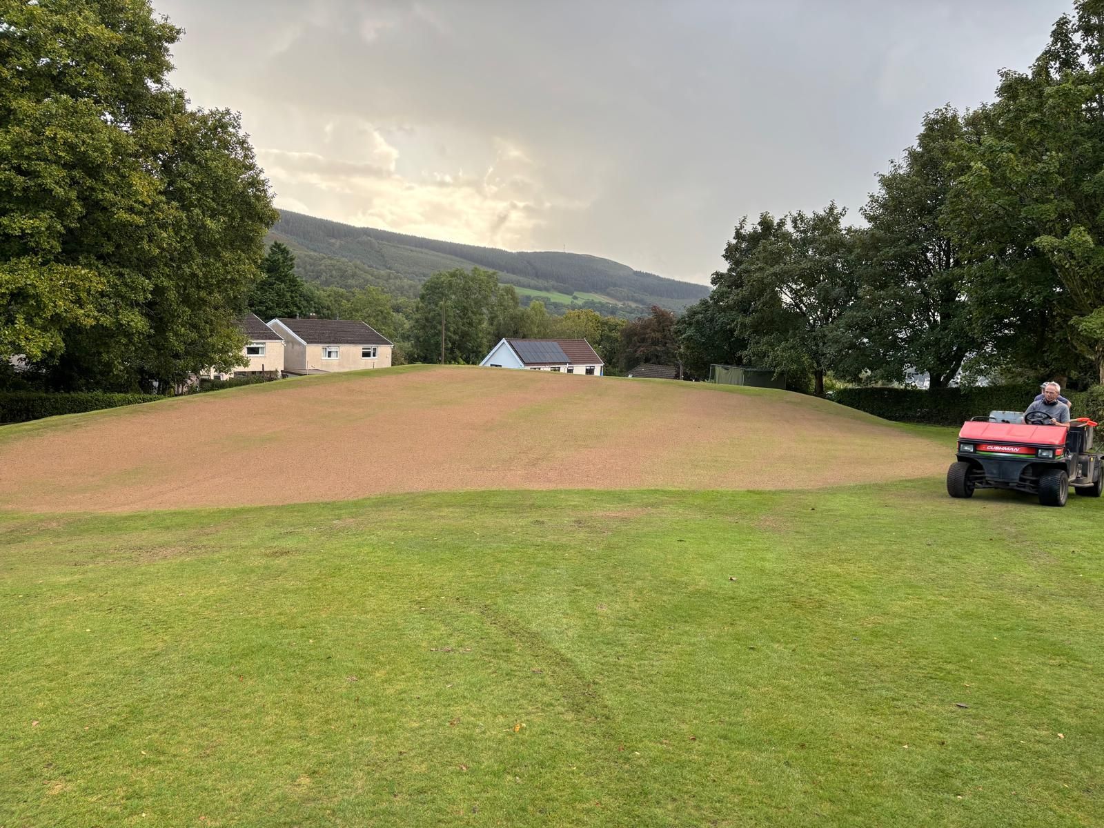 A person in a small vehicle applying red mulch on a grassy hill with houses and trees in the background.