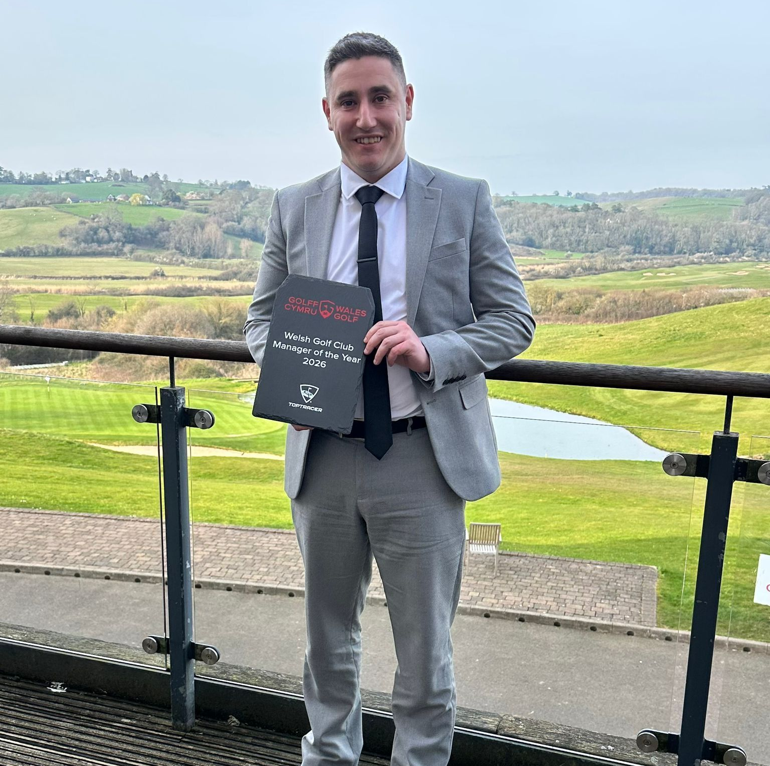 A man in a light grey suit and dark tie stands on a balcony holding a black framed award, with a green landscape behind.