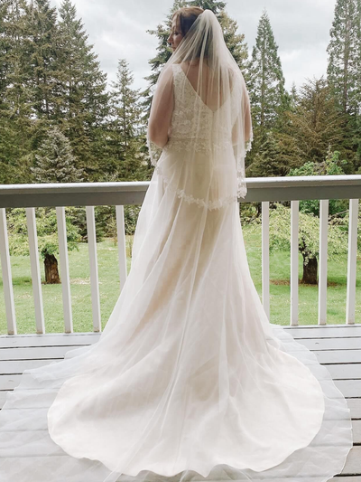 Bride in white dress raises arm, veil flowing. Holding bouquet in green field.