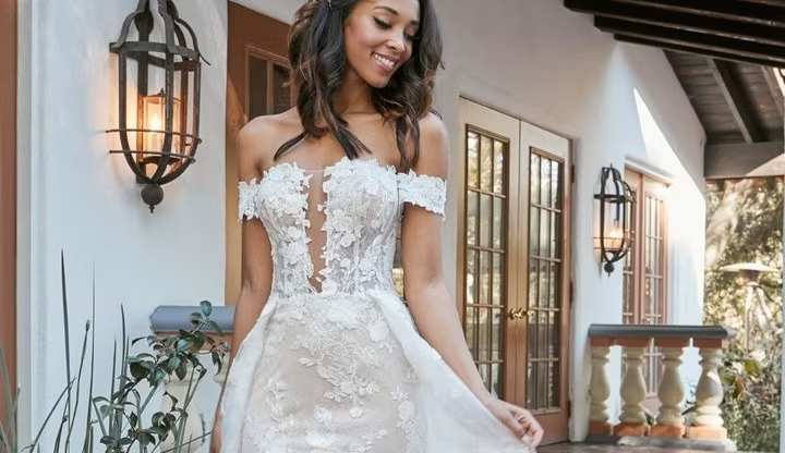Woman in off-the-shoulder lace wedding dress, smiling, standing outdoors near a building with lanterns.