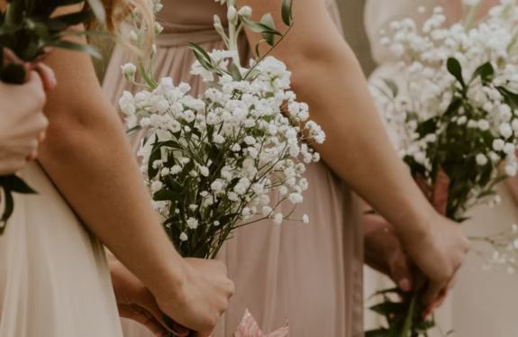 Bridesmaids in beige dresses holding bouquets of small white flowers at a wedding.