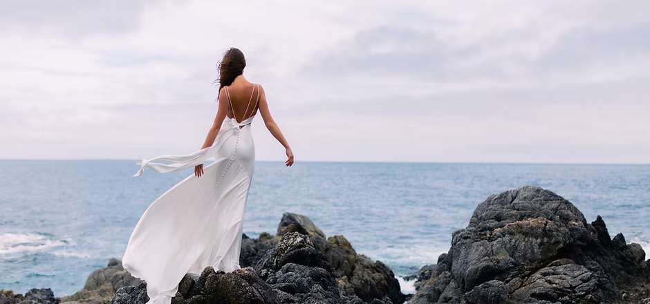 Woman in white dress standing on rocks, looking at the ocean under a cloudy sky.