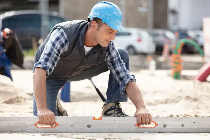 Construction worker in blue hard hat and plaid shirt crouches, gripping a concrete block at a worksite.