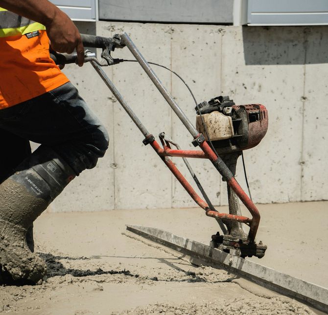 Worker using a concrete saw to cut a slab, with orange safety vest and muddy boots.