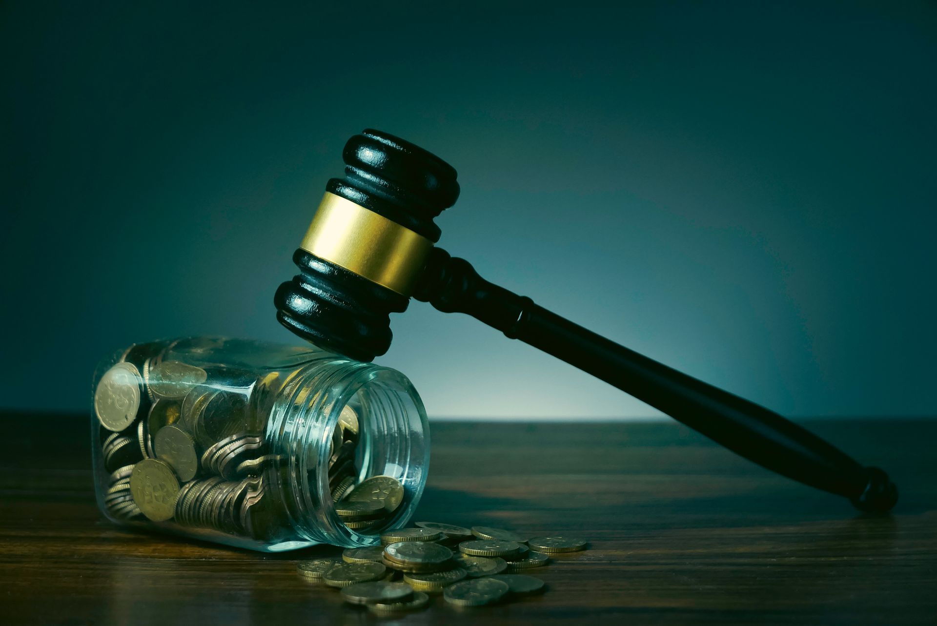 A judge’s gavel rests atop a glass jar filled with coins, with several coins spilled onto the dark wooden surface. A judge’s gavel rests atop a glass jar filled with coins, with several coins spilled onto the dark wooden surface.