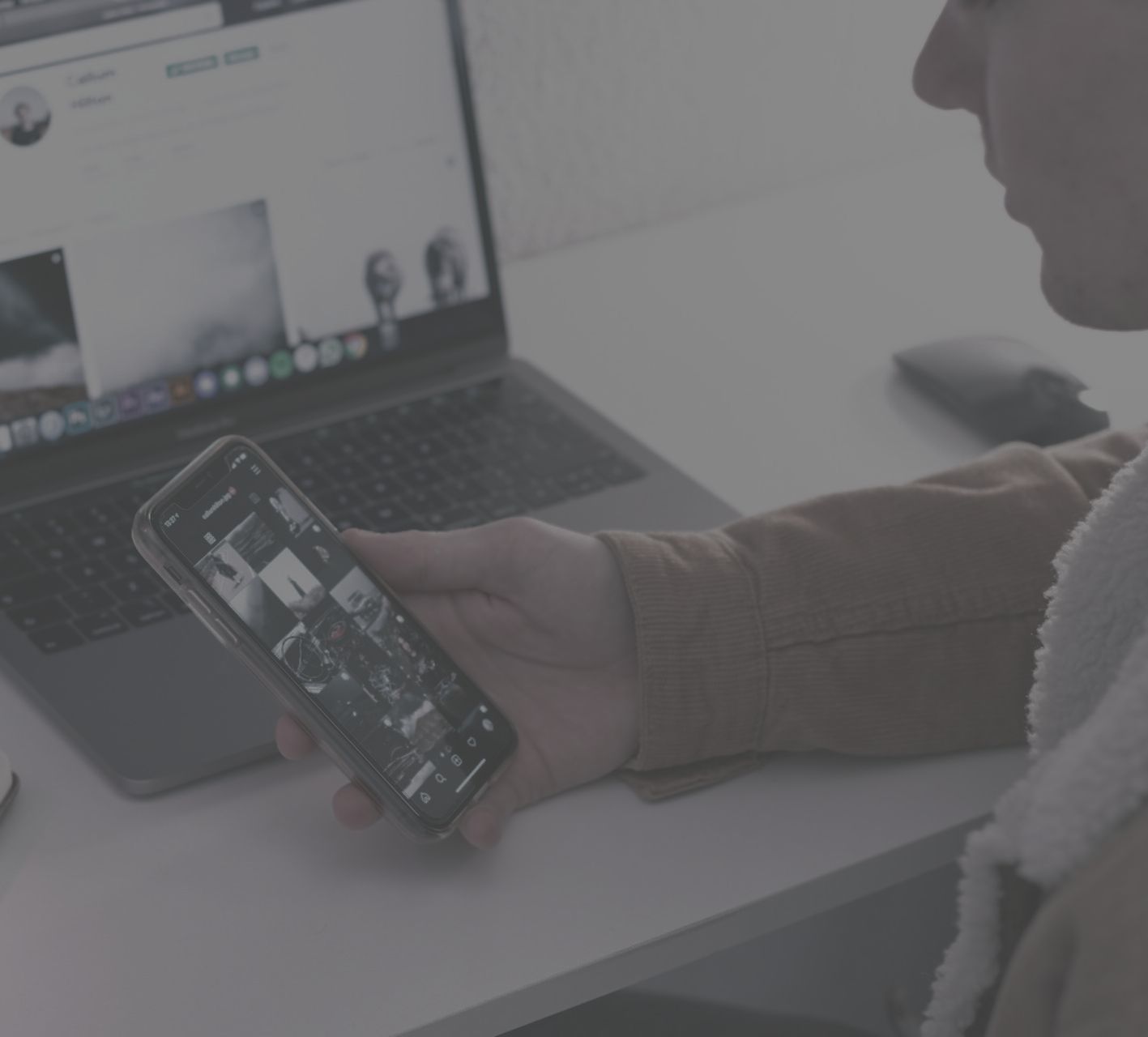 Man holding mobile phone sat at a laptop. 