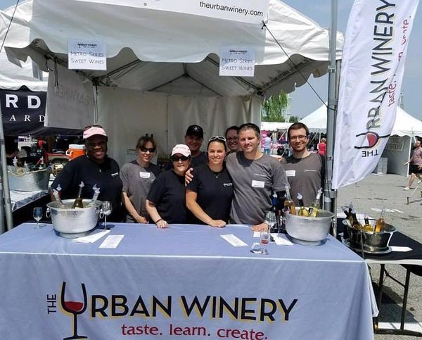 A group of people standing behind a table that says the urban winery A group of people standing behind a table that says the urban winery
