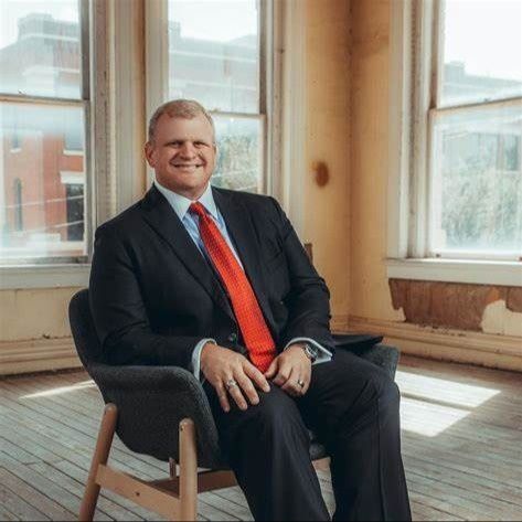 Man in a suit sits in a chair by a window, smiling. Red tie, neutral setting.