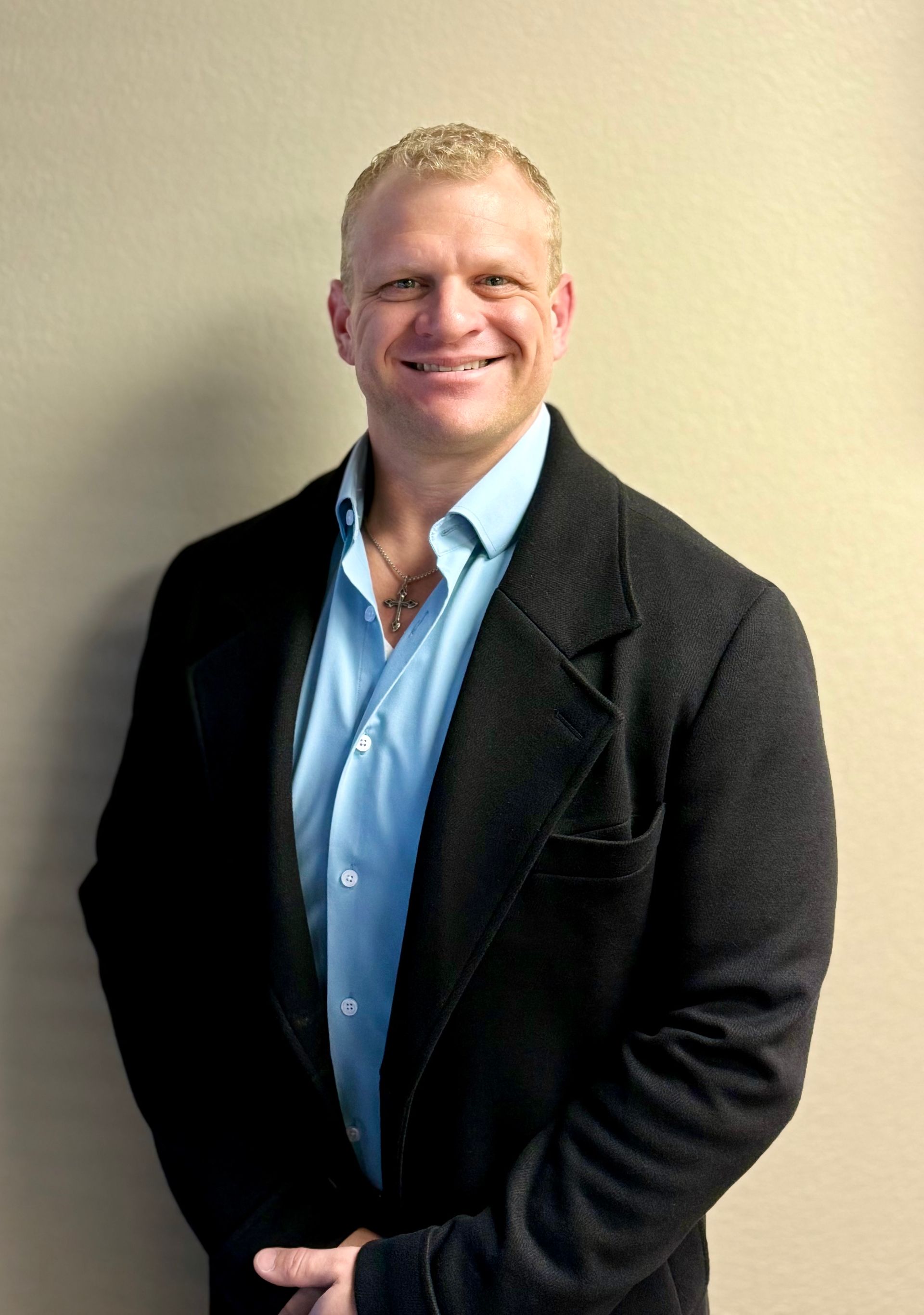 Man in a suit sits in a chair by a window, smiling. Red tie, neutral setting.
