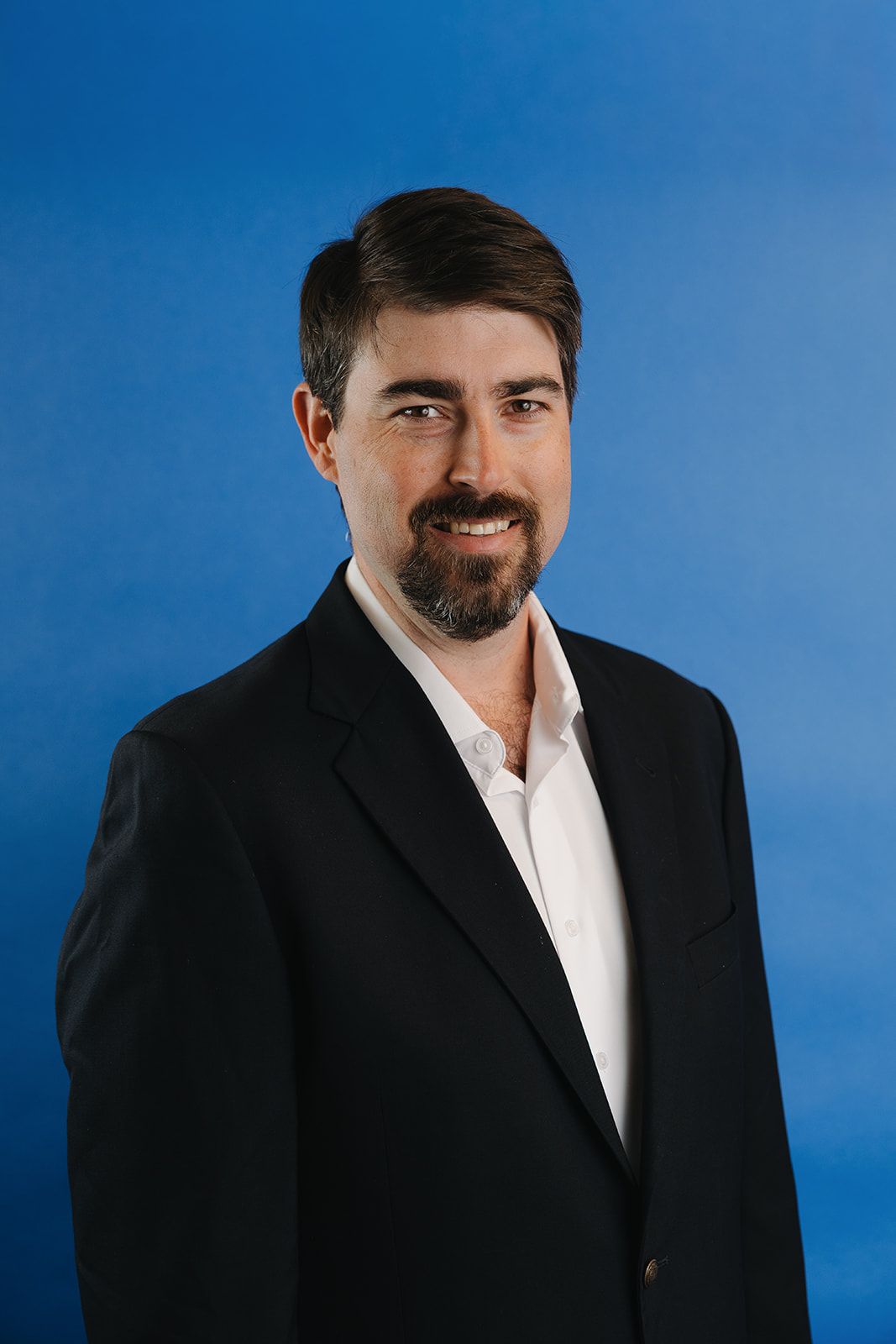 Man in blue suit sits on wooden chair, hands clasped. Dark beard, neutral expression, in industrial setting.