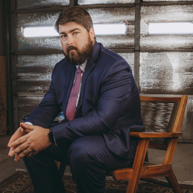 Man in blue suit sits on wooden chair, hands clasped. Dark beard, neutral expression, in industrial setting.