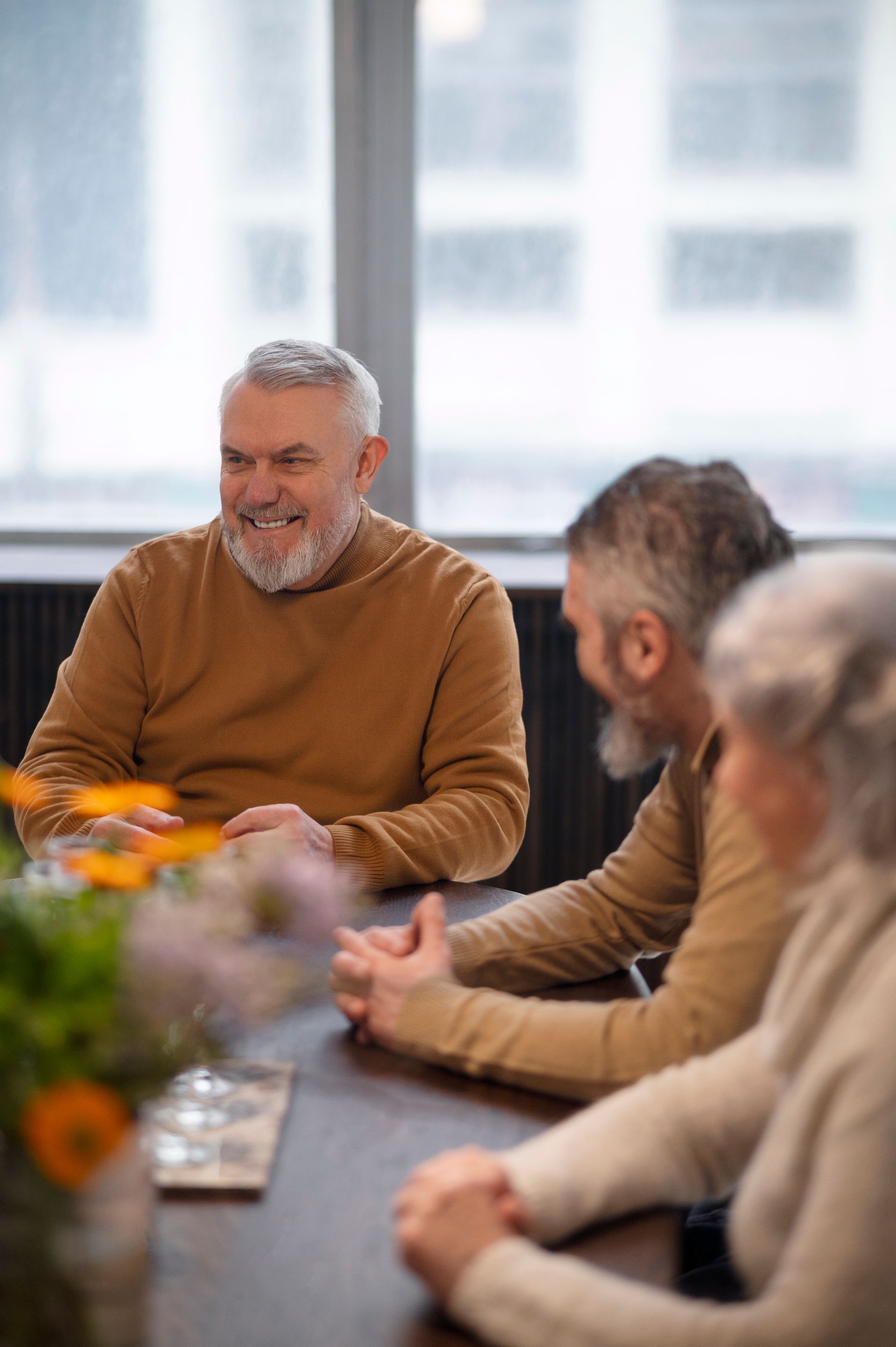 Woman, child, and older man smiling together outdoors. Sunlight, white flowers, casual clothing.