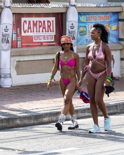 Two women in bikinis are walking down the street in front of a campari sign