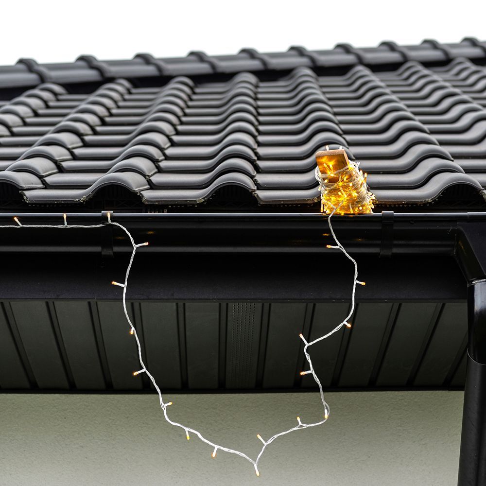 Black roof with attached gutter, lit with golden string lights. A small decorative bundle of lights sits on the roof’s edge.