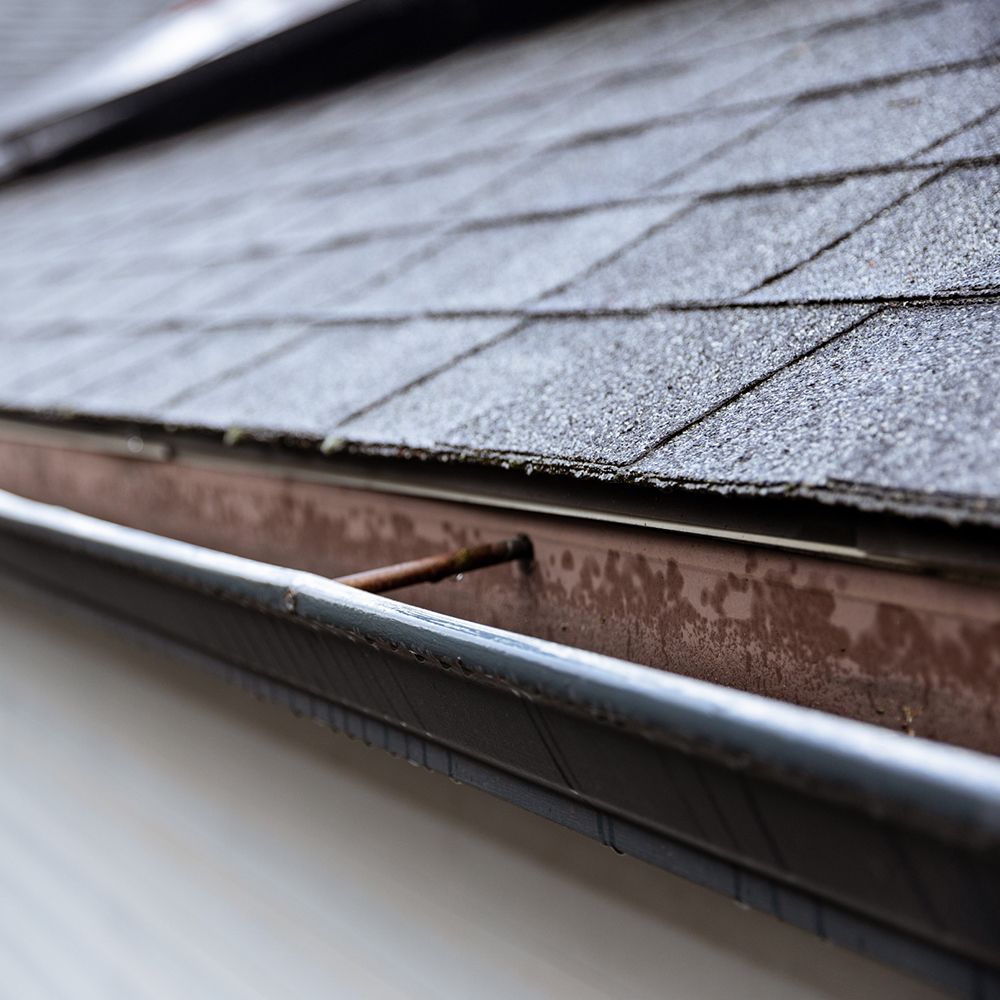 Close-up of a roof with gray shingles and a brown gutter. The gutter is partially visible with a downspout.
