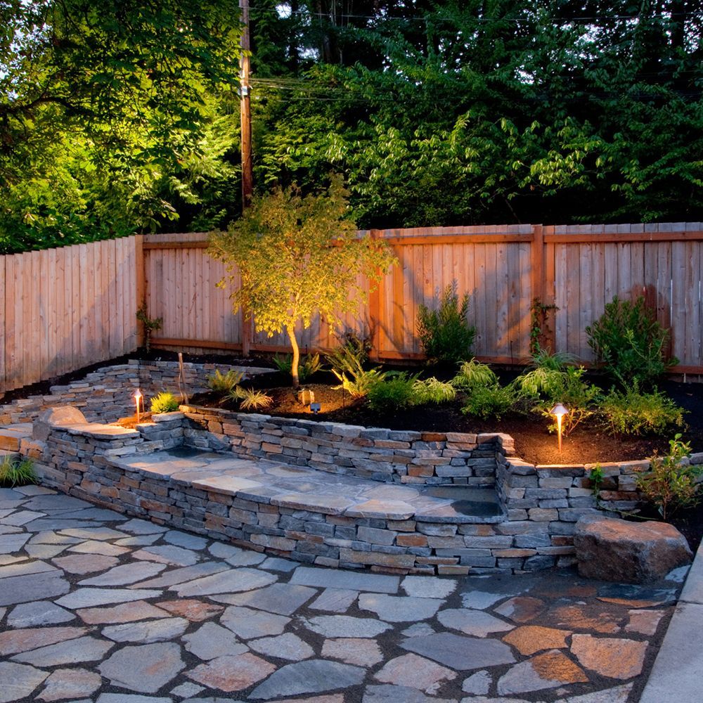 Stone patio and steps leading to a garden with a tree and illuminated by landscape lighting, wooden fence in the background.