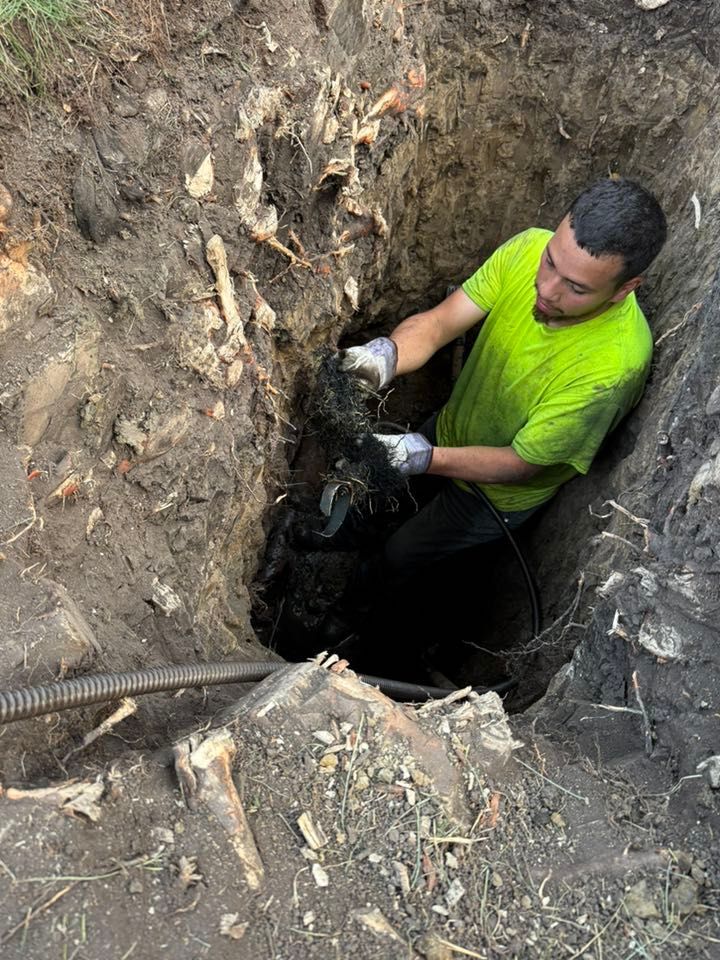 Person in blue overalls with gloves, inserting a hose into an open septic tank on a grassy lawn.