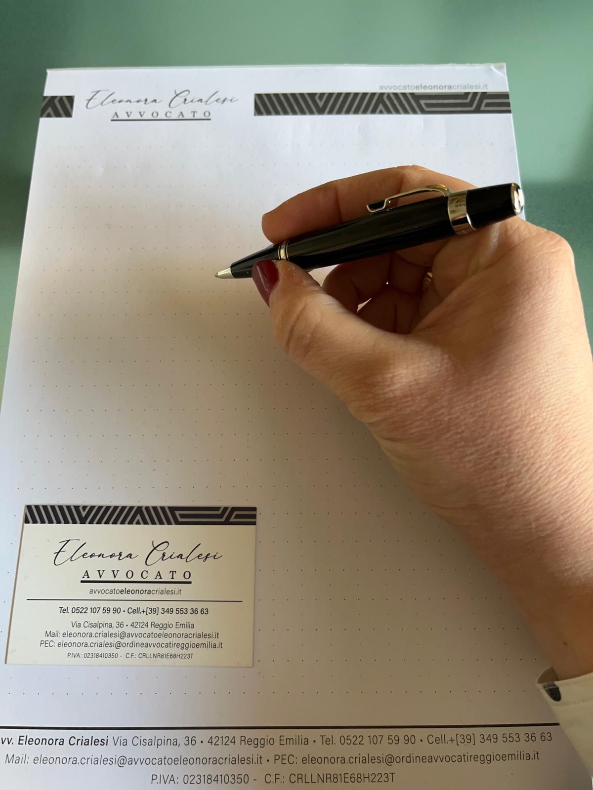 Mano di una persona che tiene una penna sopra una carta intestata con un biglietto da visita, di fronte a una parete chiara.